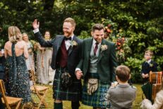 Two grooms walk down the aisle of their garden ceremony smiling as confetti is thrown. Both wear tweed jackets and kilts.