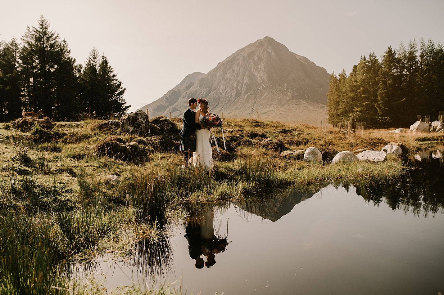 Bride and Groom hugging on grass bank à côté de l'étang avec grande montagne et arbres en arrière-plan mariages par Hayley et Craig