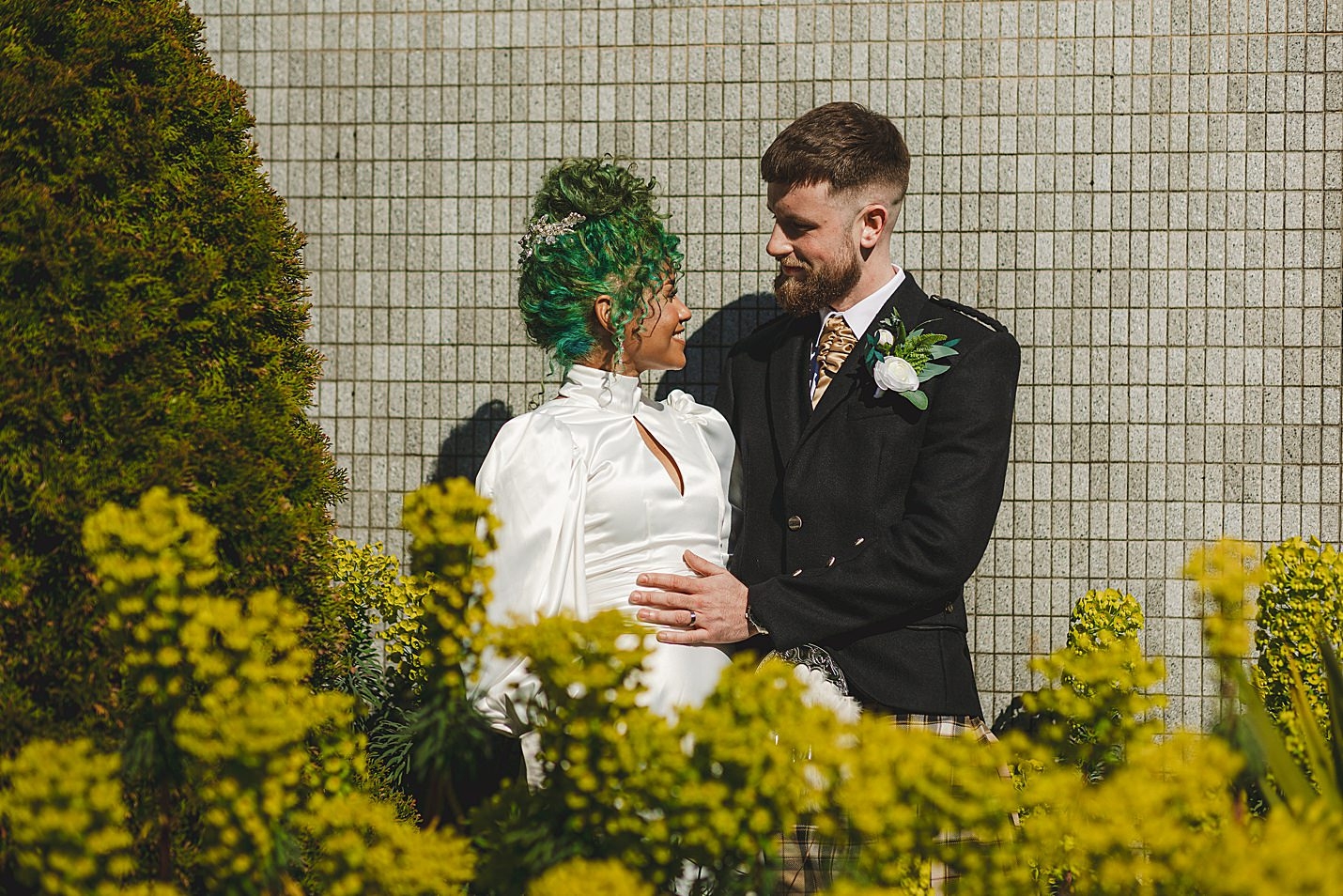 bride and groom smiling at each other in front of tiled wall with yellow plants in fore front of picture marischal college wedding clarke joss photography