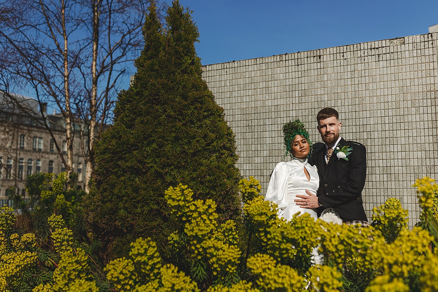 bride and groom pose to camera as they stand in front of tiled wall in gardens blue skies in background marischal college wedding clarke joss photography