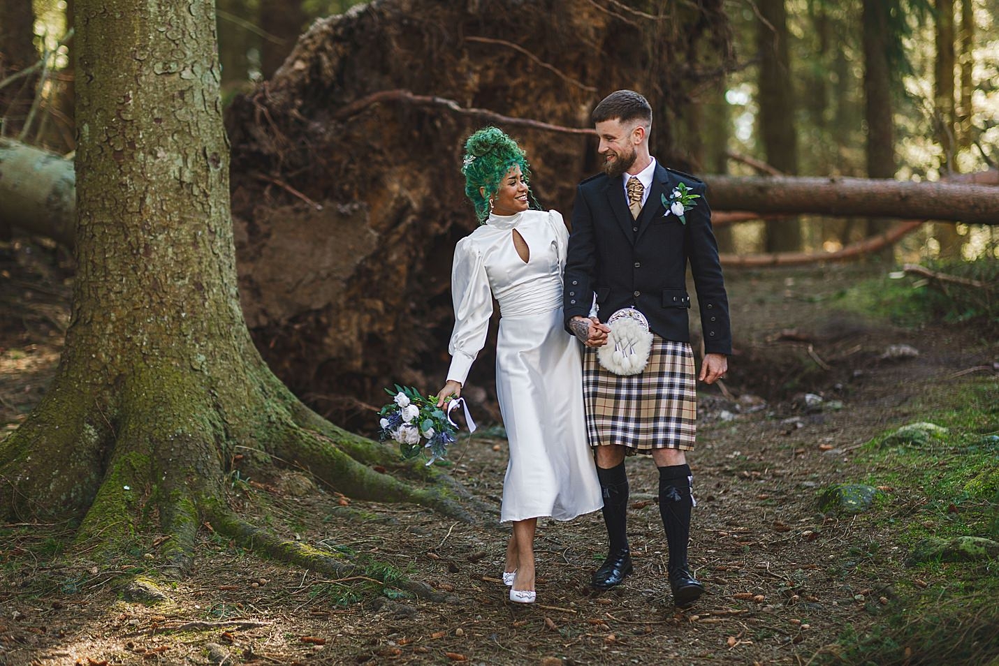bride and groom smile as they walk hand in hand towards camera through forest with fallen tree in background marischal college wedding clarke joss photography
