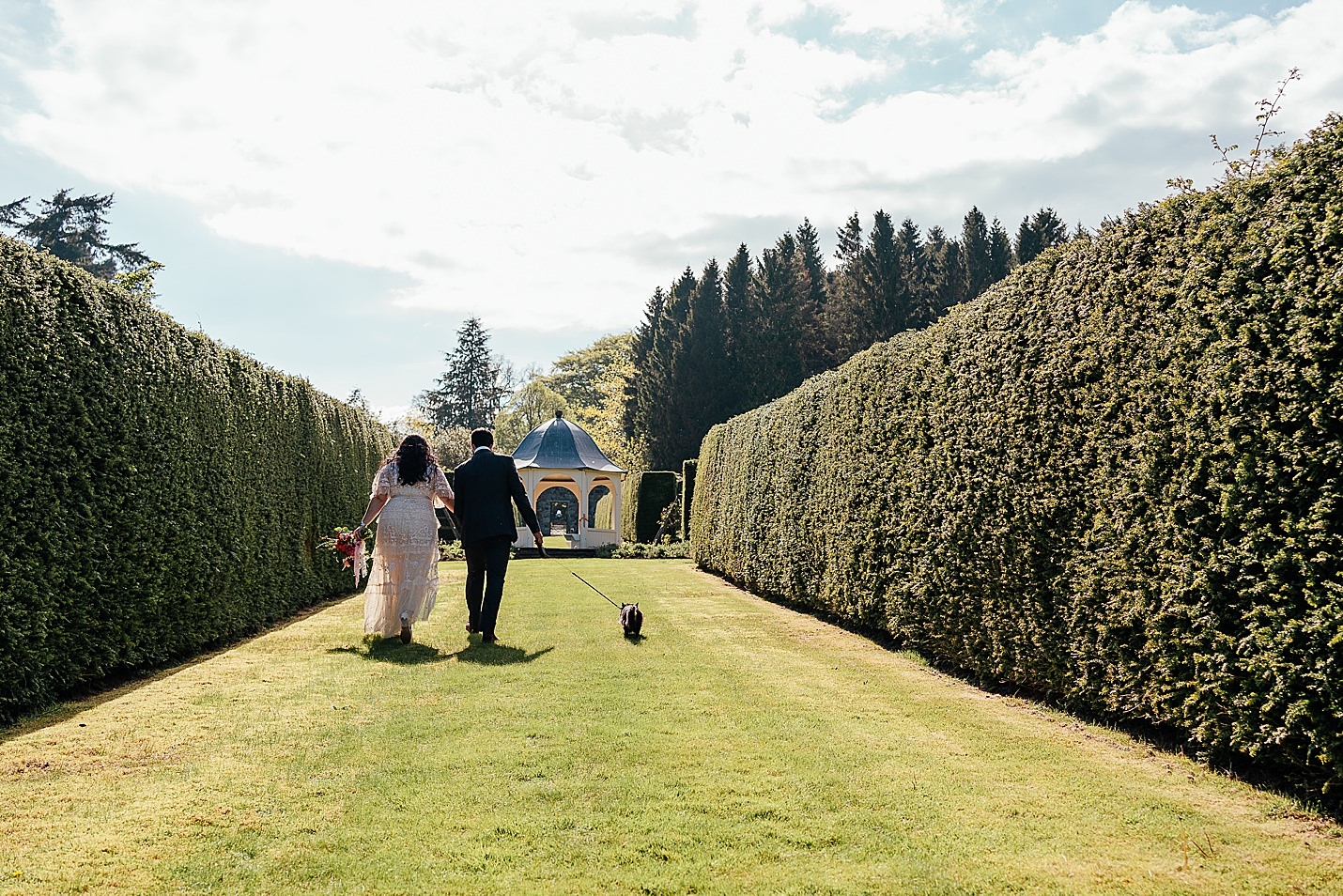bride and groom just married walking with dog in walled garden with green hedges at each side on sunny day drum castle wedding fotomaki photography