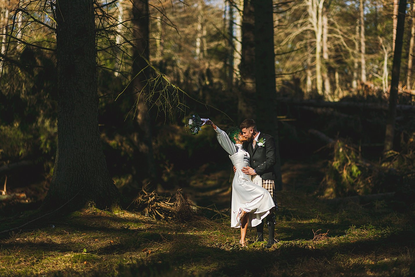 the light shines through the trees onto bride and groom as they kiss in forest clearing marischal college wedding clarke joss photography